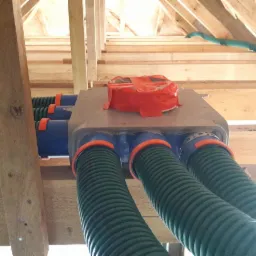 Szara skrzynka rozdzielcza rekuperacji z podłączonymi zielonymi rurami wentylacyjnymi, zabezpieczona pomarańczową taśmą, widoczna w surowym stanie poddasza z drewnianą konstrukcją więźby dachowej.