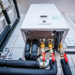 Biała jednostka wewnętrzna z wyświetlaczem i podłączonymi rurami w izolacji, widoczne zawory z czerwonymi rączkami, instalacja na białych płytkach.