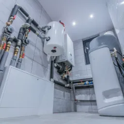 Biały zasobnik ciepłej wody użytkowej z podłączonymi rurami, zaworami i licznikiem, zamontowany w pomieszczeniu z szarymi płytkami na ścianach i podłodze, widok z dołu.