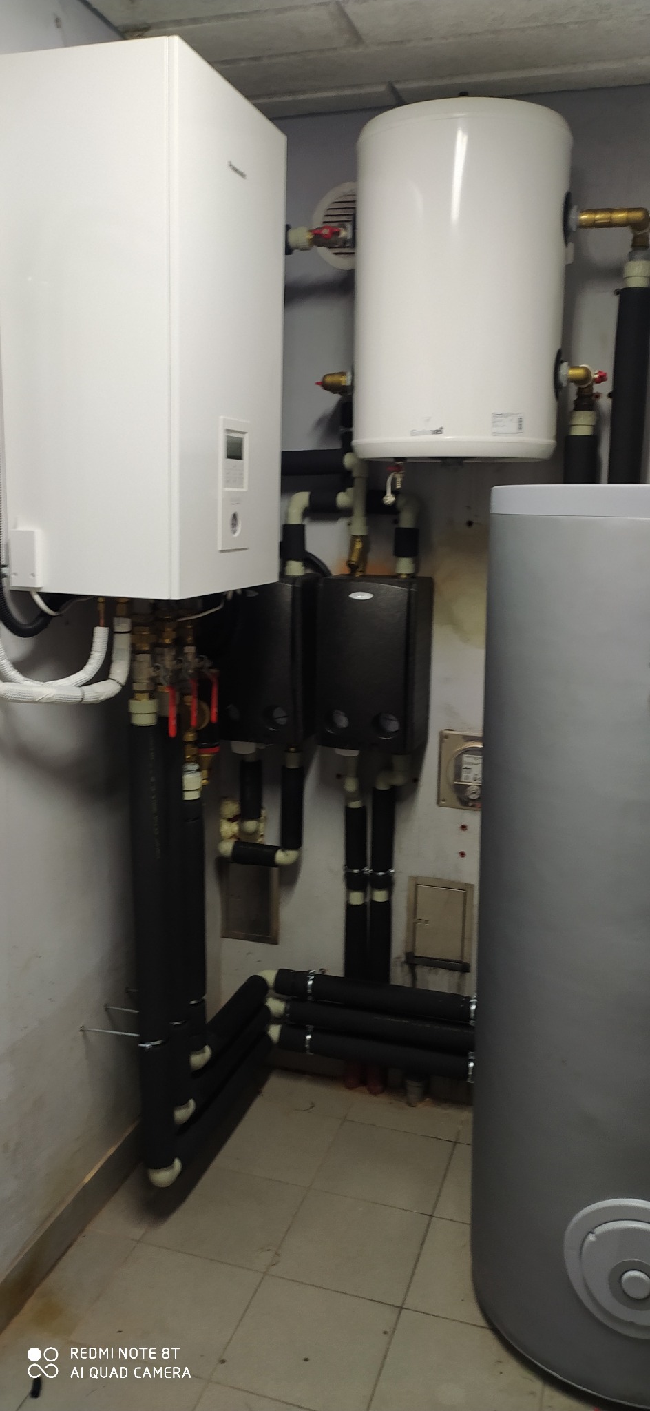 Instalacja kotłowni z białym zbiornikiem buforowym i dwoma czarnymi modułami grzewczymi, rury w izolacji z widocznymi zaworami i manometrami, płytki podłogowe w beżowym odcieniu.