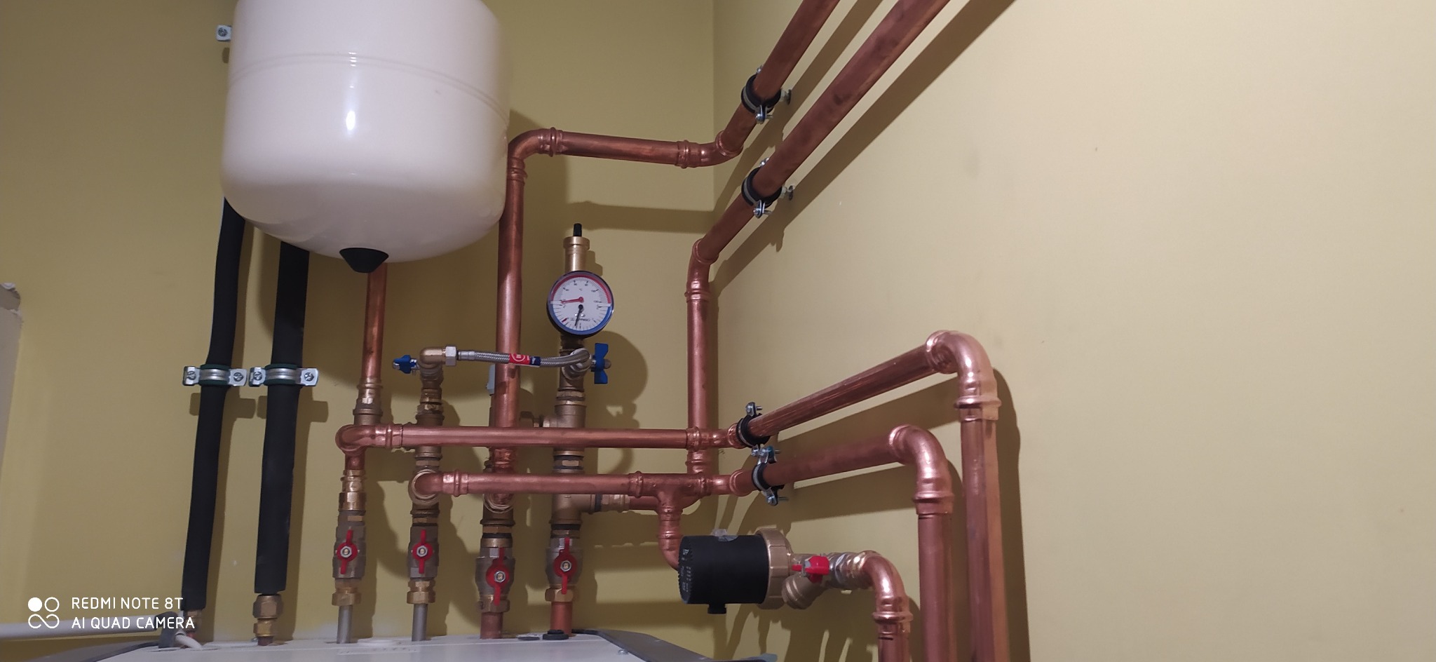 Instalacja hydrauliczna z miedzianych rur, zbiornik wyrównawczy, manometr i zawory odcinające na tle beżowej ściany.