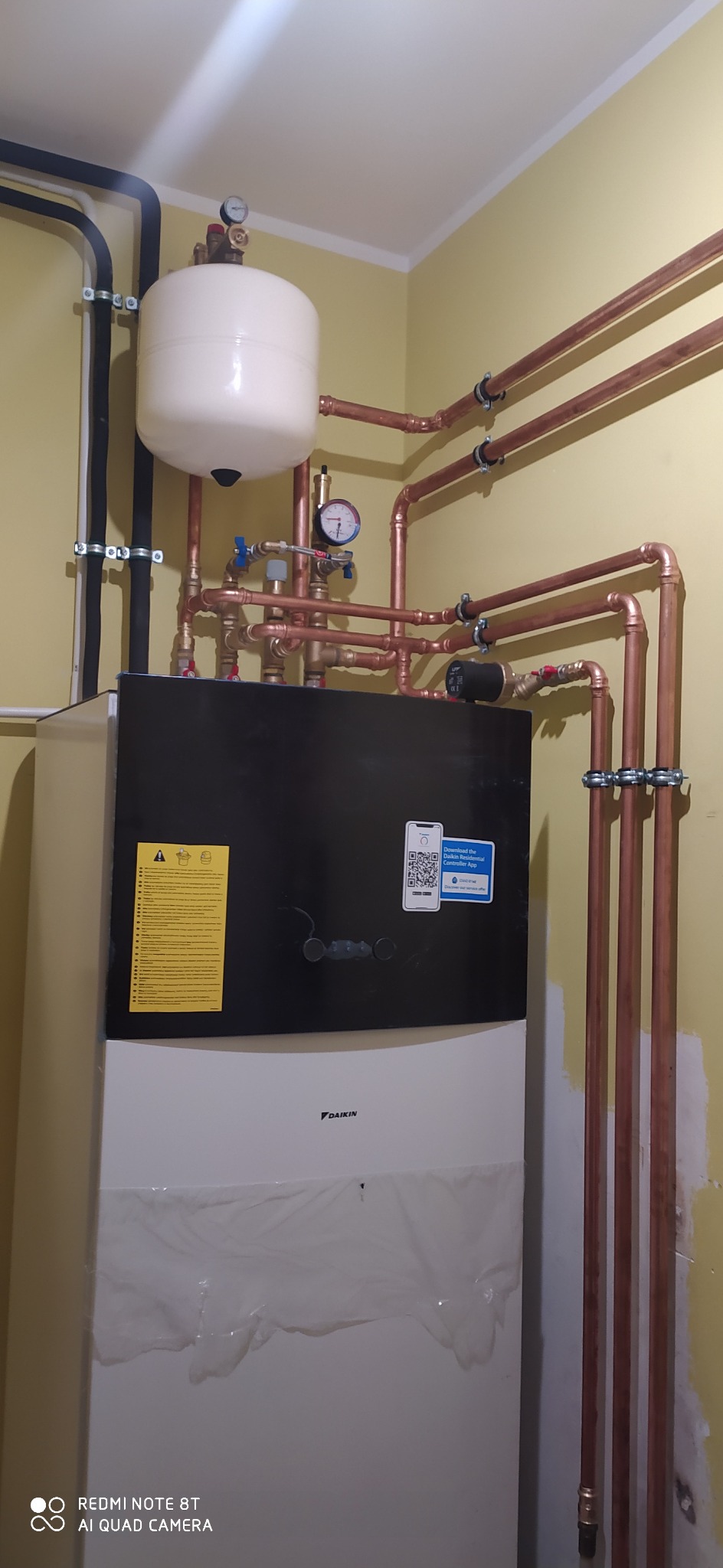 Zainstalowana jednostka Daikin z zbiornikiem wyrównawczym i miedzianą instalacją hydrauliczną, widoczne rury w uchwytach mocujących.