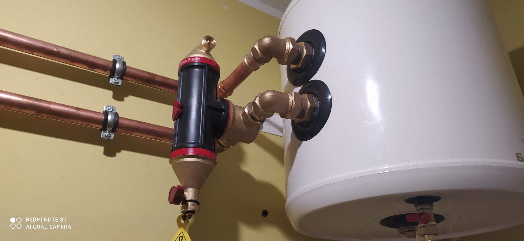 Instalacja centralnego ogrzewania: biały bojler z podłączonym filtrem magnetycznym Clean Smart, miedziane rury przymocowane do żółtej ściany.