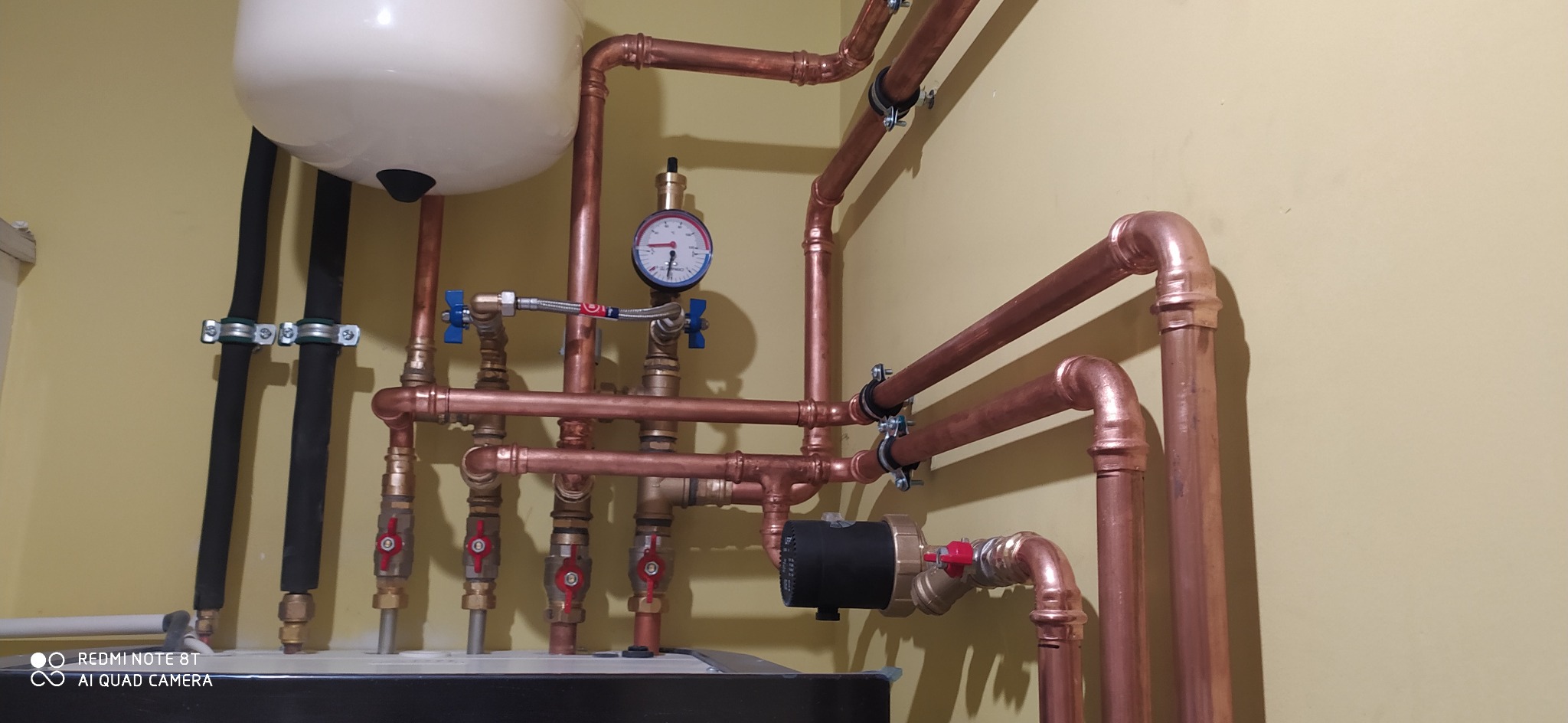Kompleksowa instalacja hydrauliczna z miedzianych rur, widoczny manometr, zawory kulowe z czerwonymi i niebieskimi uchwytami oraz pompa obiegowa.