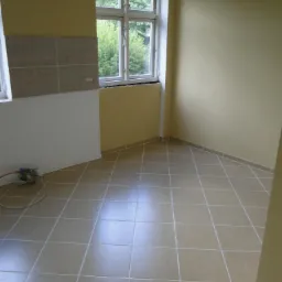 Wykończona podłoga z płytek w jasnym odcieniu, widoczne fragmenty ścian w odcieniach beżu i bieli, okno z widokiem na zieleń. Fragment instalacji przy ścianie.