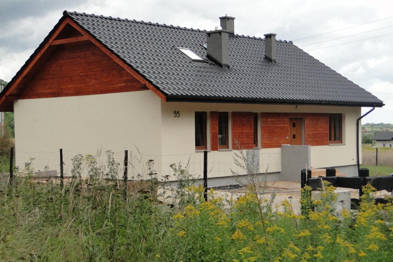 Nowoczesny dom jednorodzinny z ciemną dachówką, drewnianym szczytem i jasną elewacją. Widoczne okna z drewnianymi okiennicami i numer domu '55'.