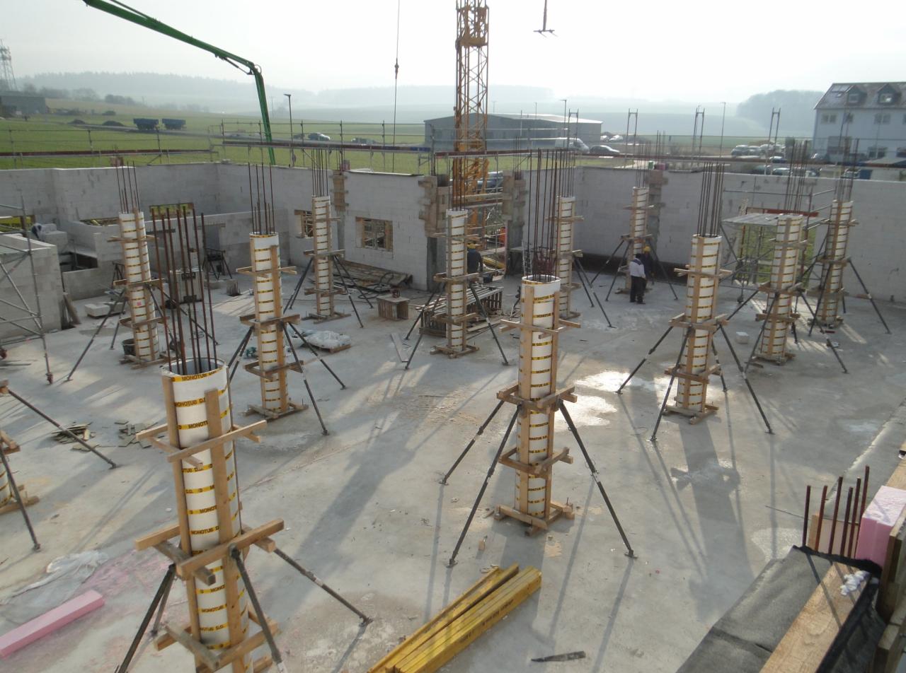 Budowa domu: Wylane fundamenty z kolumnami zbrojeniowymi zabezpieczonymi szalunkiem, widoczne ściany fundamentowe, dźwig budowlany w tle.