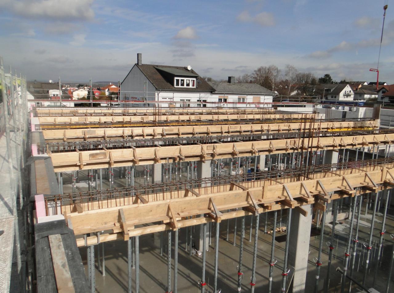 Wysoki kąt ujęcia szalunku stropu zbrojonego na podporach stalowych, w tle budynki mieszkalne pod błękitnym niebem.