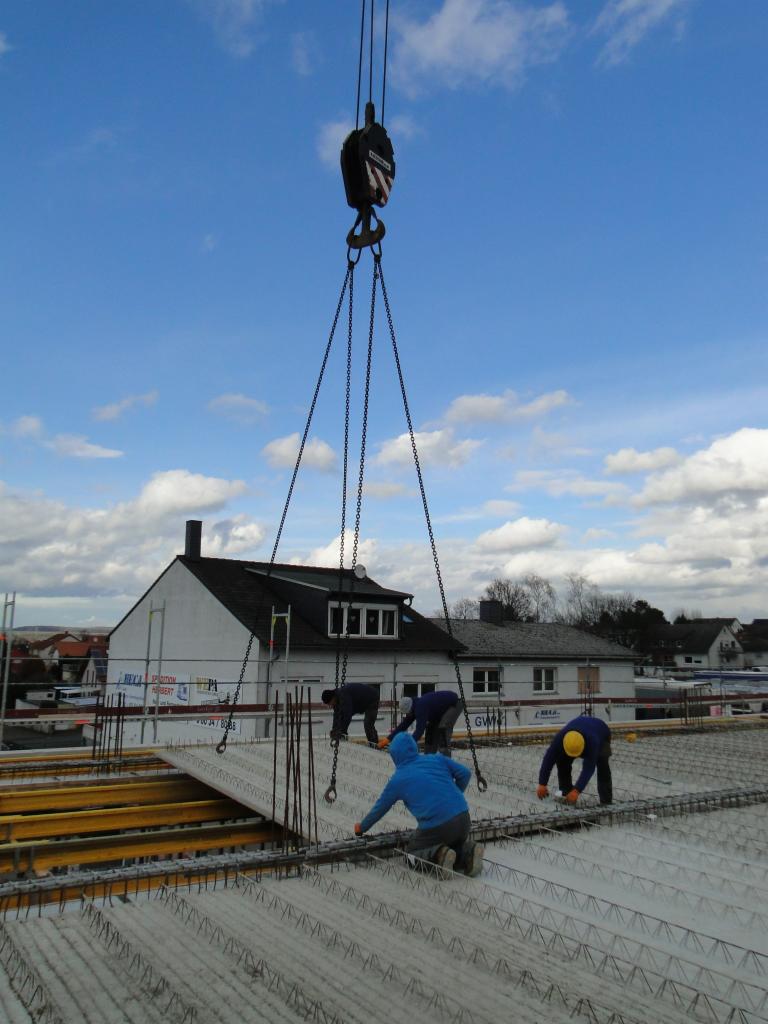 Robotnicy montują zbrojenie stropu na budowie domu, płyta prefabrykowana zawieszona na haku dźwigu, w tle biały budynek z poddaszem.