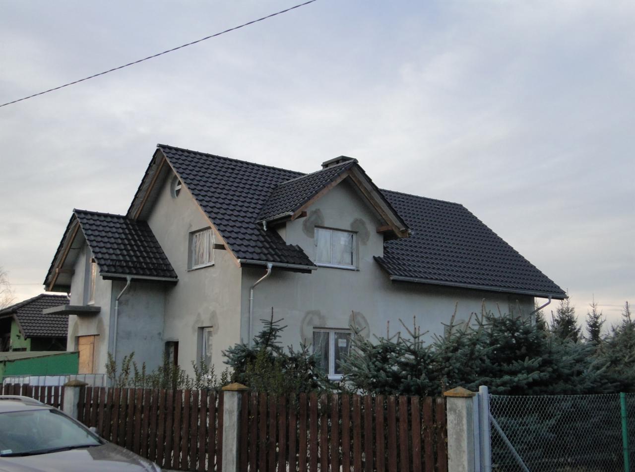 Dom dwukondygnacyjny z ciemną dachówką, w trakcie prac elewacyjnych, widoczny zza drewnianego płotu i iglaków, z szarą elewacją i oknami zabezpieczonymi folią.