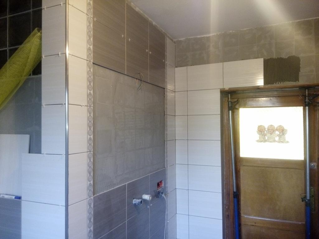 Ściana w trakcie układania płytek ceramicznych w dwóch odcieniach szarości, widoczne krzyżyki dystansowe, fragment drzwi z naklejką aniołków oraz żółta folia ochronna w rogu.