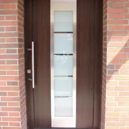 Brązowe drzwi wejściowe z matowym przeszkleniem i srebrnymi listwami, zamontowane w murze z cegły. Długi, srebrny uchwyt po lewej stronie.