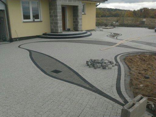 Ułożona kostka brukowa w oryginalny wzór przed domem z szarą elewacją i kamiennym wejściem. Na powierzchni leżą jeszcze kostki do ułożenia.