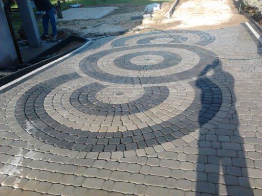 Ułożona kostka brukowa tworząca wzór koncentrycznych okręgów w odcieniach szarości. Widoczny cień osoby fotografującej.