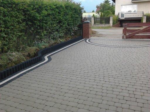 Ułożona szara kostka brukowa na podjeździe z dekoracyjnymi obramowaniami i żywopłotem w tle. Widoczny fragment bramy i domu.