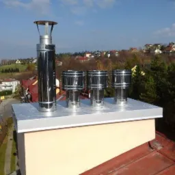 Instalacja wkładu kominowego ze stali nierdzewnej na dachu budynku, z widokiem na okoliczne domy i zieleń. Cztery elementy wkładu, jeden wysoki i trzy niższe.