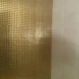 Ściana wykończona złotą mozaiką z małych kwadratowych płytek przylegająca do ściany wyłożonej białymi, błyszczącymi płytkami w łazience.