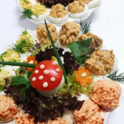 Dwa talerze z dekoracyjnie podanymi jajkami faszerowanymi, z różnymi nadzieniami i ozdobami z warzyw, w tym jajko-muchomor z pomidora i majonezu.