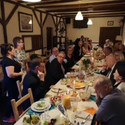 Sala restauracyjna z drewnianymi belkami stropowymi, oświetlona wiszącymi lampami, gdzie goście siedzą przy długim stole zastawionym potrawami i napojami podczas uroczystości.