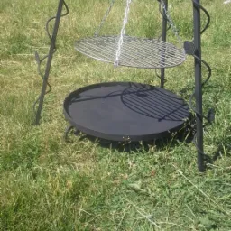 Kuty trójnóg z dwoma poziomami: okrągłą płytą paleniskową i rusztem na łańcuchu, ustawiony na trawie.