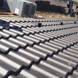 Wadim Syczewski M Kwadrat - Częściowo pokryty dach z czarnej dachówki ceramicznej z elementami wentylacyjnymi, widoczne drewniane łaty i paczki dachówek w tle, praca w trakcie wykonywania.