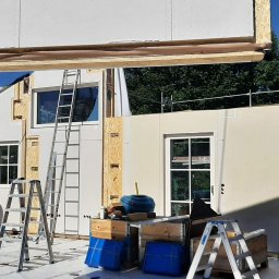 Wadim Syczewski M Kwadrat - Montaż prefabrykowanych ścian domu szkieletowego, widoczny pracownik na drabinie w kapeluszu, elementy konstrukcyjne podnoszone na miejsce, narzędzia i materiały budowlane na podłodze, częściowo...