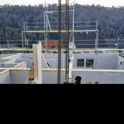 Wadim Syczewski M Kwadrat - Wznoszenie konstrukcji domu z prefabrykatów na tle zalesionego wzgórza, widoczne rusztowania i łańcuchy dźwigu podczas montażu ścian.