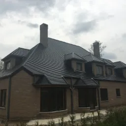 Nowo wybudowany dom z cegły z ciemną dachówką i kominem, otoczony zielenią, na tle pochmurnego nieba.