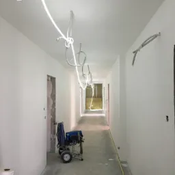 Korytarz w trakcie prac wykończeniowych, ściany świeżo pomalowane na biało, widoczny agregat malarski, rolka papieru i przewody elektryczne pod sufitem.