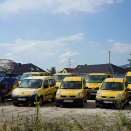 Flota firmowa: kilka żółtych samochodów dostawczych z napisem 'Wylewki' zaparkowanych na placu, w tle budynki i błękitne niebo.