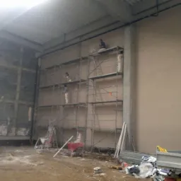 Praca tynkarska na rusztowaniu w hali: ekipa nakłada tynk na ścianę. Część ściany surowa, część otynkowana. Widoczne narzędzia i maszyna do tynkowania.