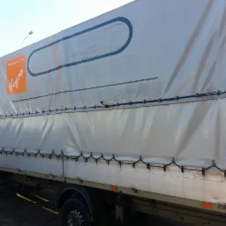 Biały samochód dostawczy z plandeką, na której widnieje pomarańczowe logo 'Vegas' i napis 'ROLETY', zaparkowany na kostce brukowej, z budynkiem mieszkalnym w tle.