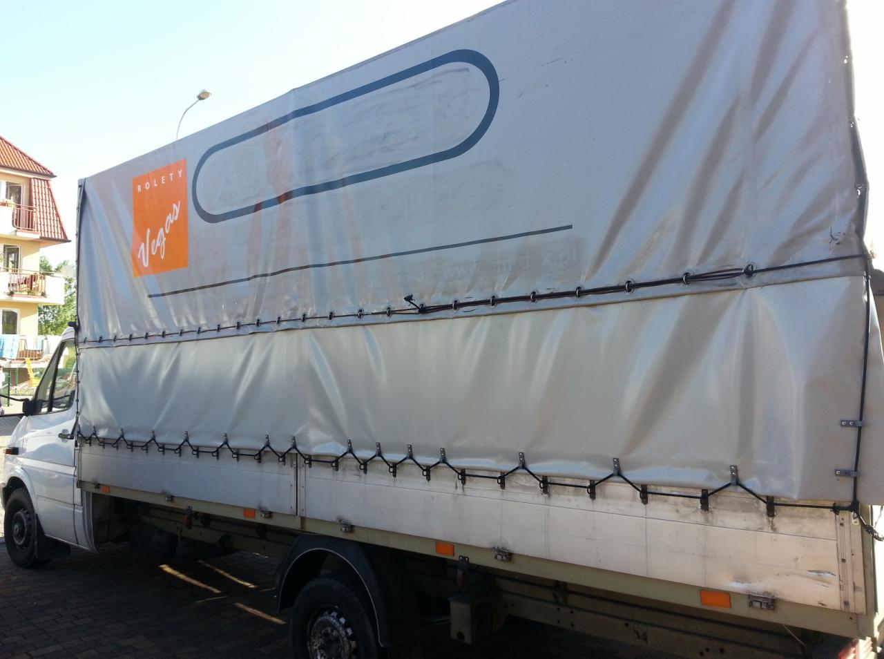 Biały samochód dostawczy z plandeką, na której widnieje pomarańczowe logo 'Vegas' i napis 'ROLETY', zaparkowany na kostce brukowej, z budynkiem mieszkalnym w tle.