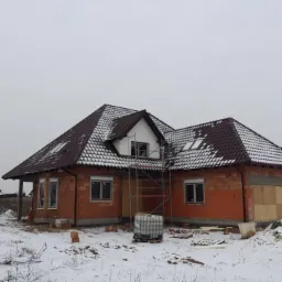 Dom w trakcie budowy, z brązowym dachem pokrytym częściowo śniegiem, czerwona cegła, szare niebo, rusztowanie przy oknie, zimowy krajobraz.