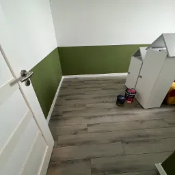 Pomieszczenie po malowaniu ścian na biało i oliwkowo zielono, widoczne dwie puszki farby Śnieżka i szafki na podłodze z paneli.