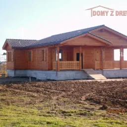 Gotowy dom z bali z ciemną dachówką, widok z zewnątrz. Na pierwszym planie ziemia, w tle winnica i sylwetka osoby na drabinie. Widoczne logo firmy.