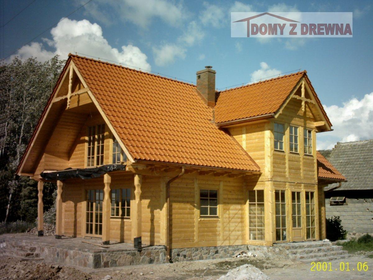 Jasny, dwukondygnacyjny dom z bali z pomarańczową dachówką ceramiczną, widoczny komin i fragment starego budynku w tle. Surowy stan.