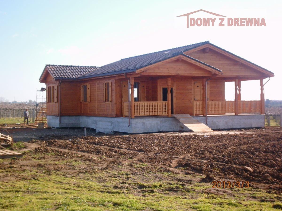 Gotowy dom z bali z ciemną dachówką, widok z zewnątrz. Na pierwszym planie ziemia, w tle winnica i sylwetka osoby na drabinie. Widoczne logo firmy.