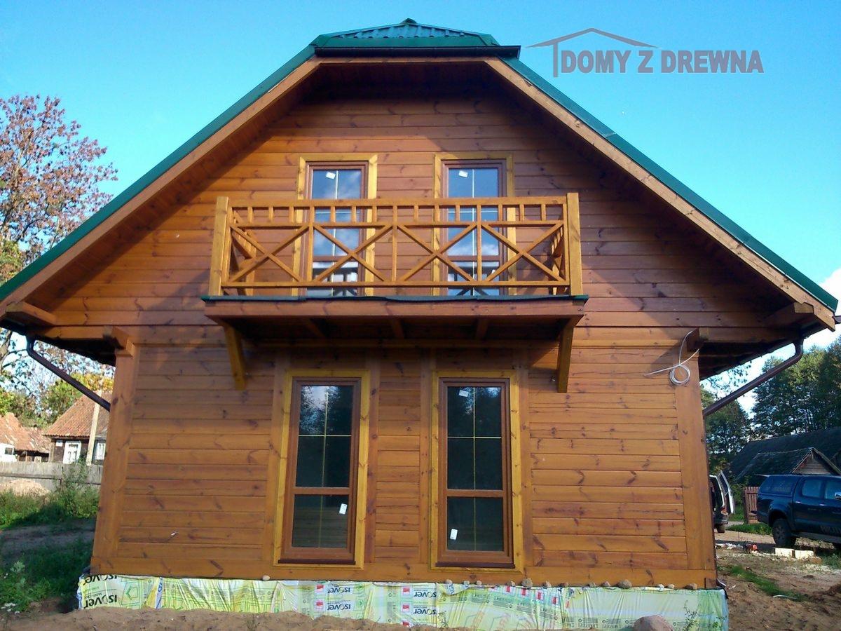 Dwupiętrowy dom z bali z balkonem, wykończony na brązowo, z zielonym dachem na tle błękitnego nieba. Widoczne okna i fragment terenu wokół budynku.