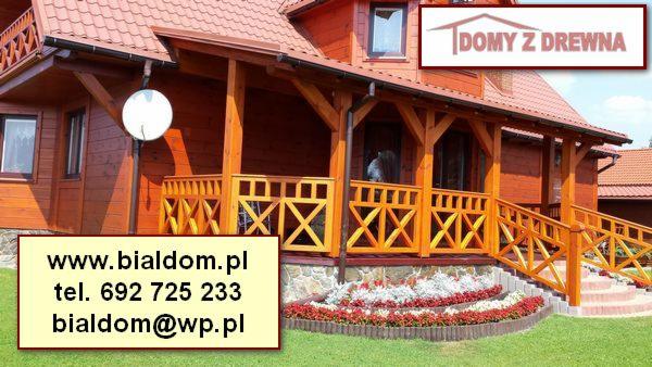 Drewniany dom z czerwonym dachem, widoczny z zewnątrz z dekoracyjnym balkonem, kwiatami i anteną satelitarną. Dom z bali.