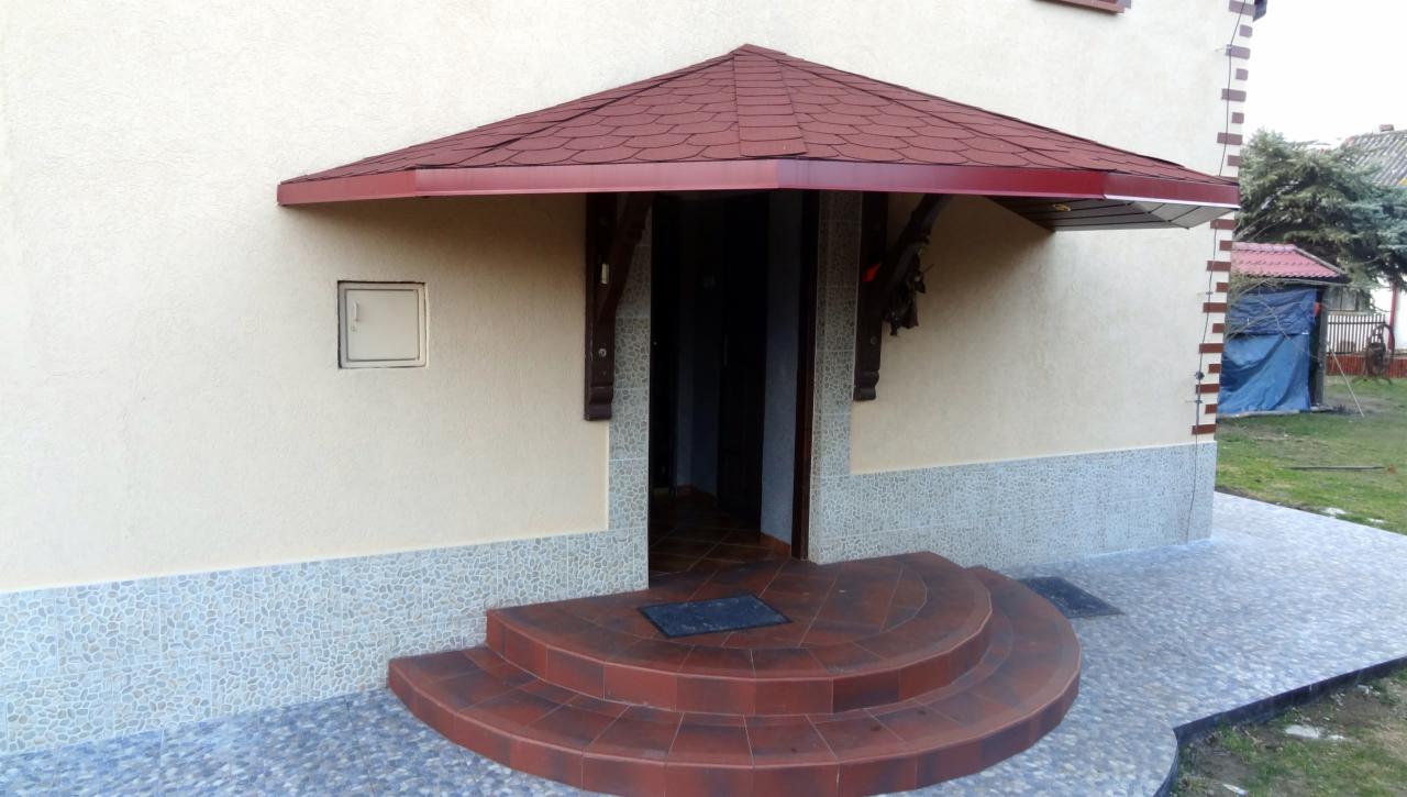 Wejście do domu z czerwonym zadaszeniem nad drzwiami, wykończone dekoracyjnym tynkiem i mozaiką. Schody wejściowe wyłożone czerwoną terakotą. Widoczny fragment elewacji.