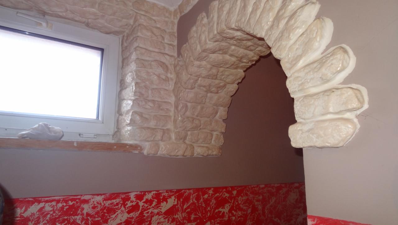 Dekoracyjny łuk z imitacji kamienia wokół okna i przejścia, kremowa kolorystyka, czerwone zdobienia na dole ściany, nierówne wykończenie, surowy styl.