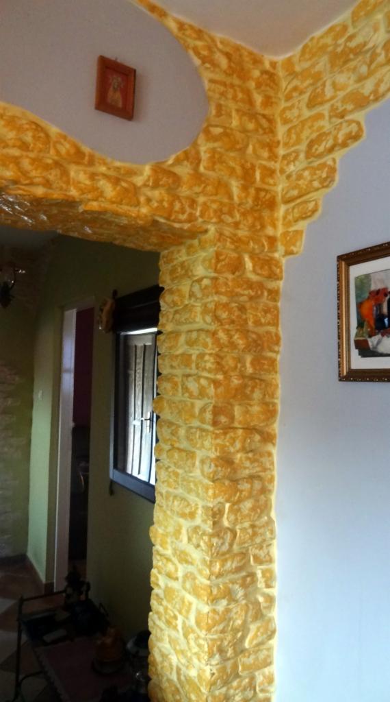 Żółta kolumna z efektem kamienia wewnątrz budynku, dekoracyjna faktura imitująca cegły, widoczne obrazy na ścianach i fragmenty pomieszczeń.