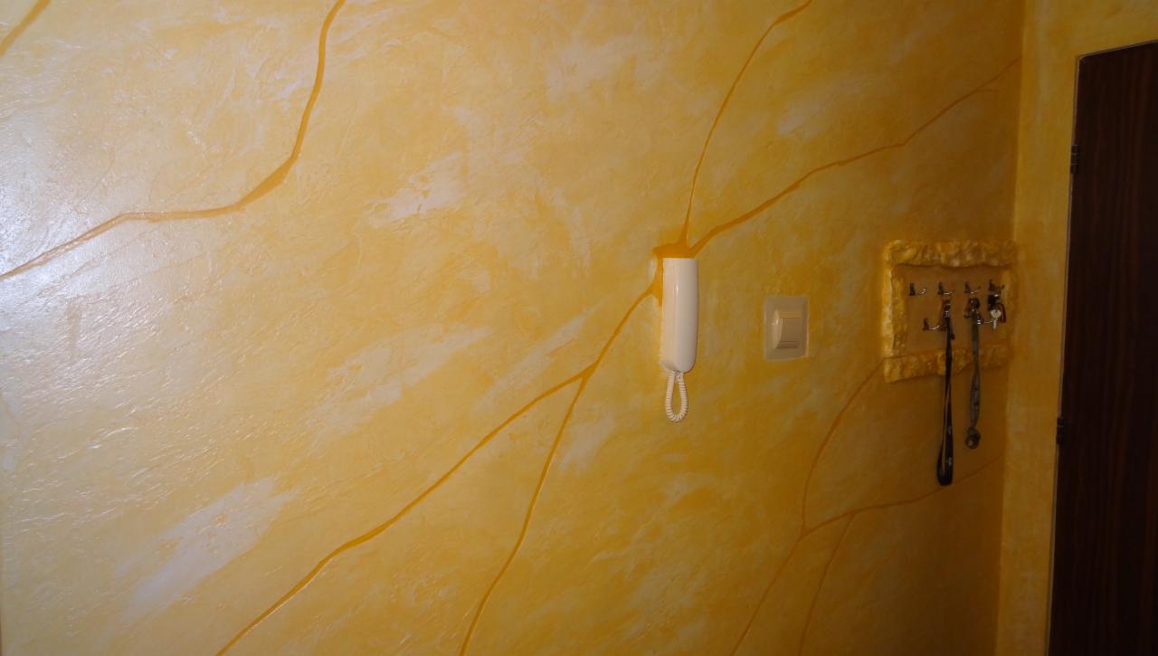 Żółta ściana z dekoracyjnym wzorem przypominającym żyły kamienia, z zamontowanym domofonem i wieszakiem na klucze. Widoczny fragment drzwi.