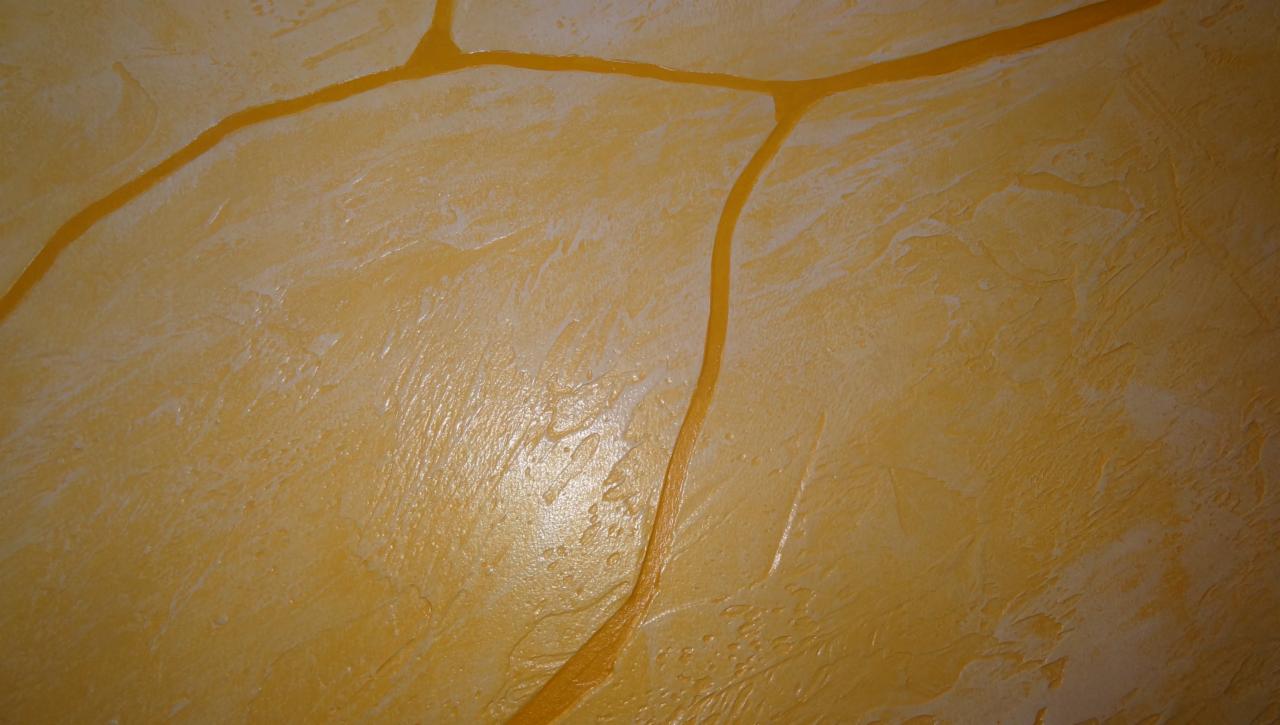 Żółta, dekoracyjna struktura tynku imitująca kamień z wyraźnymi, ciemniejszymi spoinami. Widoczne subtelne nierówności i refleksy światła.