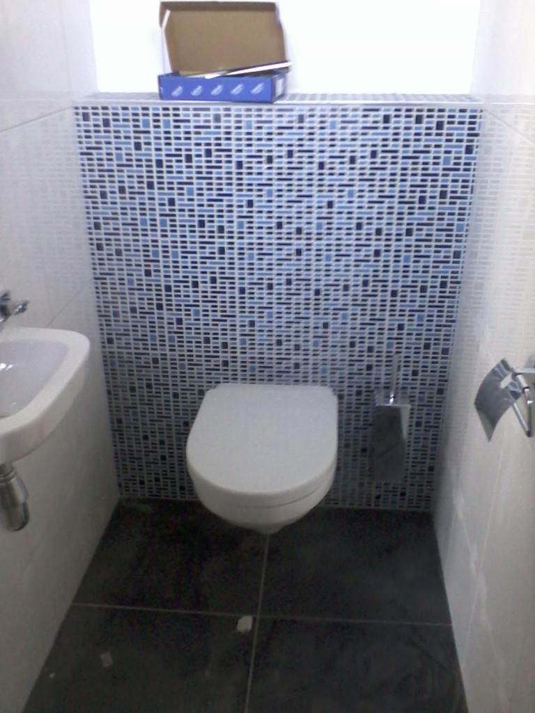 Wykończona toaleta z mozaiką w odcieniach błękitu za sedesem i ciemną podłogą. Umywalka po lewej stronie, akcesoria łazienkowe po prawej. Na górze karton z akcesoriami.