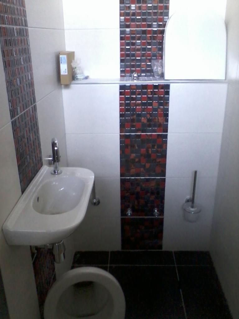 Wykończona łazienka z białą umywalką i toaletą, czarne płytki podłogowe. Ściany wyłożone białymi i mozaikowymi płytkami w odcieniach czerwieni i czerni.