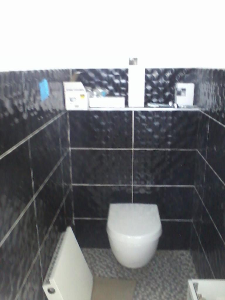 Nowoczesna toaleta z czarnymi, błyszczącymi płytkami i szarą podłogą z mozaiki. Wisząca muszla klozetowa oraz półka z akcesoriami dopełniają minimalistyczny wystrój.