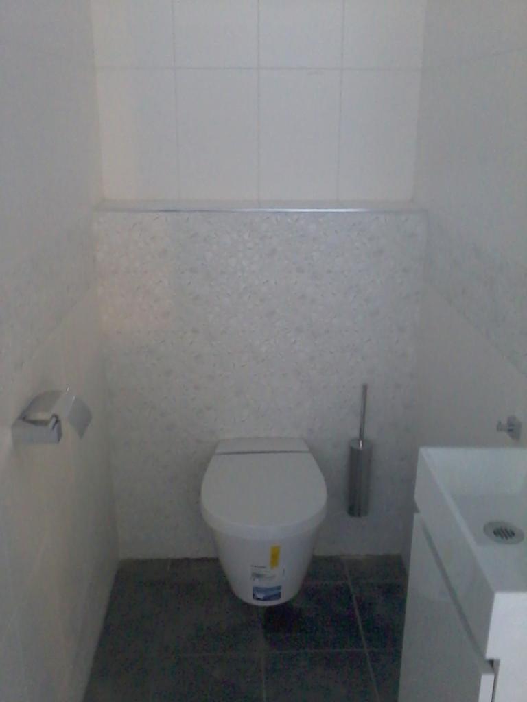 Wykończona, mała łazienka z białą toaletą, umywalką i wzorzystymi płytkami ściennymi. Widoczne detale: uchwyt na papier, szczotka do WC. Minimalistyczny design.