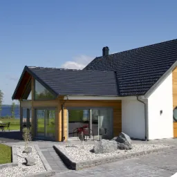 Nowoczesny dom jednorodzinny z ciemną dachówką ceramiczną, drewnianą elewacją i dużymi przeszkleniami w słoneczny dzień.